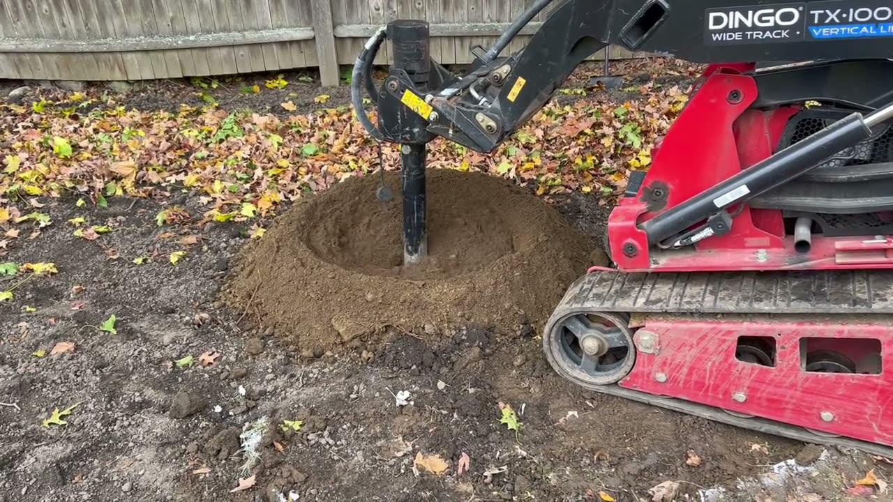 A red mini skid steer running an auger into the ground of a fenced back yard, soil mounding up around the bit